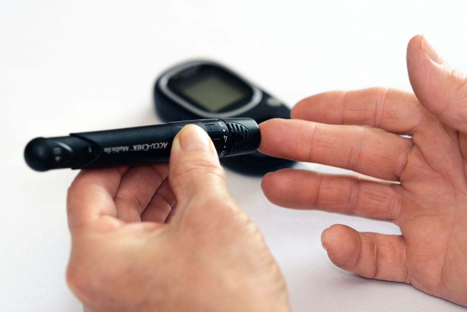 Close-up of hands using a lancet device to check blood sugar with a glucose meter in the background