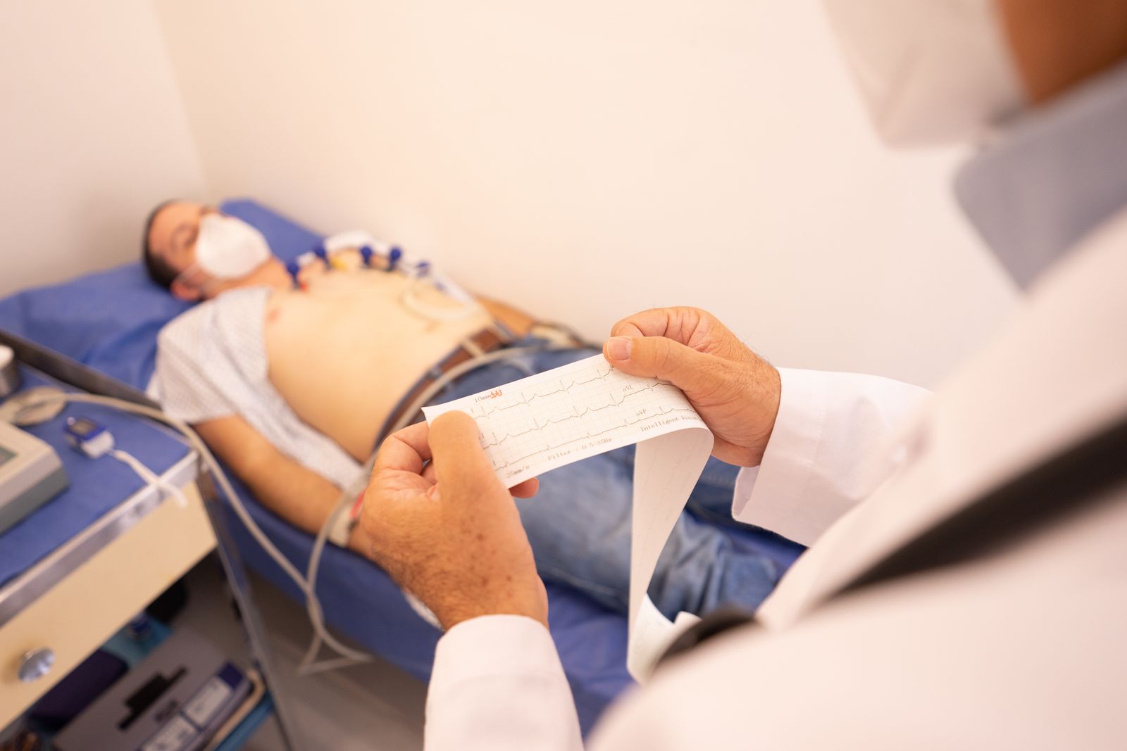 A physician reviewing an EKG paper strip while a patient rests on the exam table with heart-monitor leads attached to the chest
